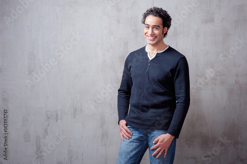 Charming and self-confident. Handsome young arabic man holding hand in pockets and looking at camera while standing against grey background © luengo_ua