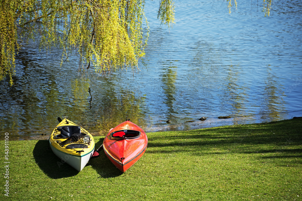 Two kayaks lie under a tree in the grass on the shore of a lake, ready for leisure activity, copy space in the blue water