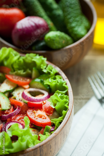 Rustic salad of fresh tomatoes, cucumbers, red onions and lettuce, dressed with olive oil and ground pepper in a wooden bowl. Top view