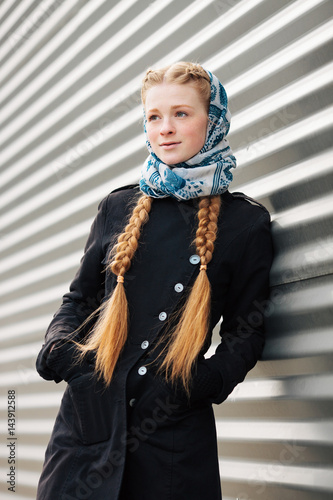 Young beautiful fashionable redhead woman with braids hairdo in blue white headcraft stylish denim black trench jacket posing outdoors against urban style background of metal strips
