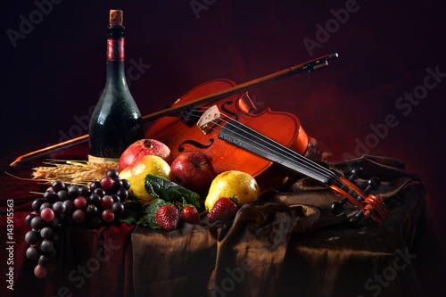 Violin with a bow on a red background next to a bottle of old wine and wet fruit