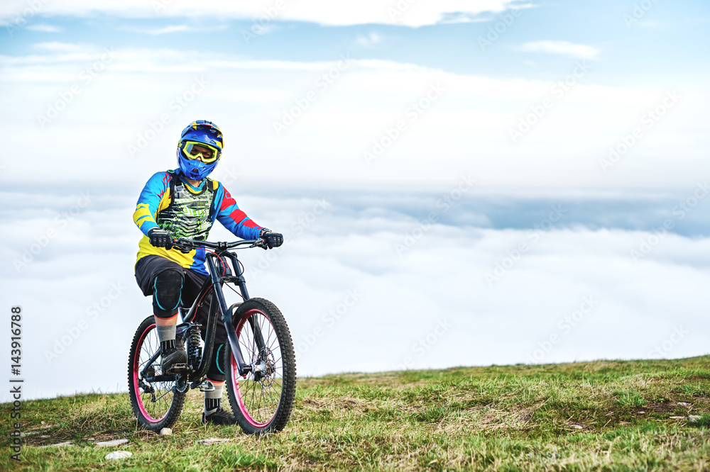 A young guy sits on a mountain bike atop a mountain, when below the mountains low clouds A young guy sits on a mountain bike atop a mountain, when below the mountains low clouds