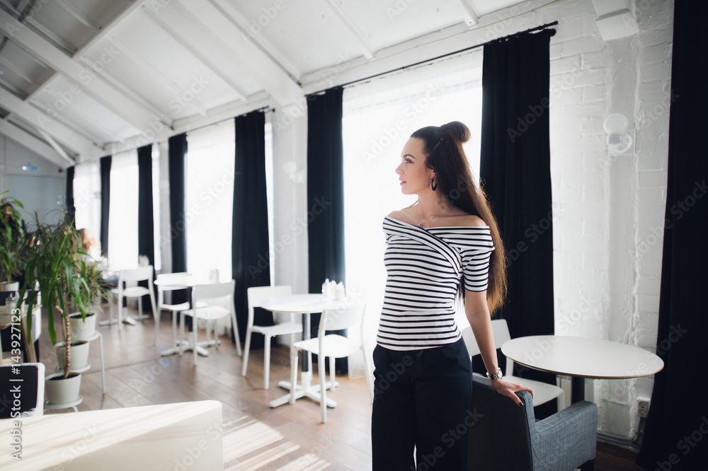 Young charming female is dreaming about something good, while is standing in a cafe. Thoughtful woman is looking away and waiting for a call on cell telephone.