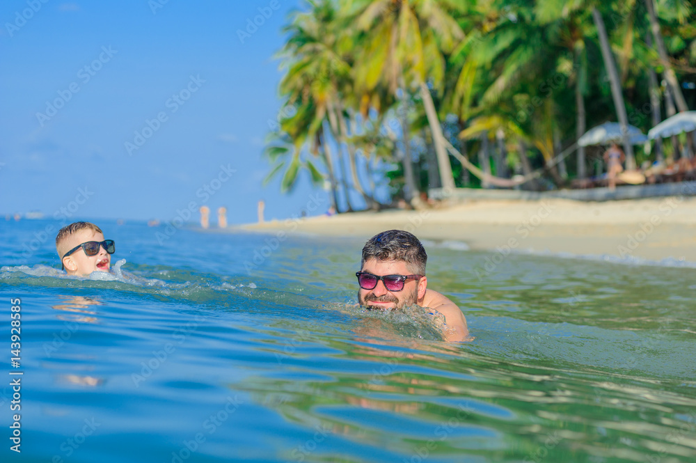 Happiness portrait in tropical water: blond boy lying on the water surface as a crocodile and his brown-haired bearded father swimming. Both in sunglasses, playing catch-up - dad wins