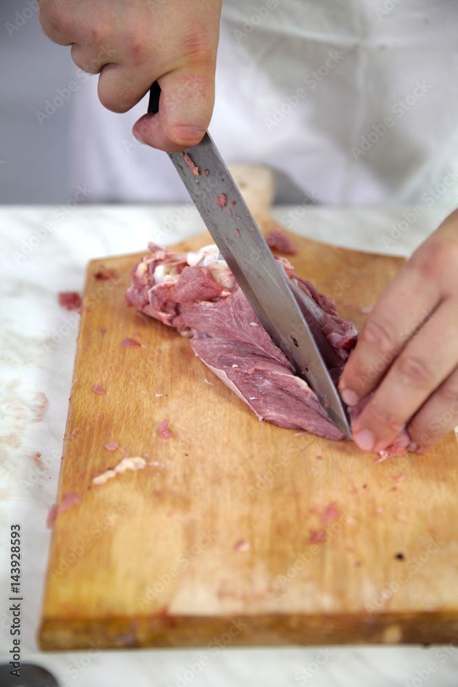 A chef is carefully cutting meatinto smaller pieces on a wooden board. He is preparing dinner for the guests.