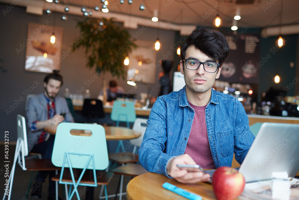Fototapeta premium Portrait of handsome young man wearing glasses and jeans jacket looking at camera while working with laptop and smartphone at table in cafe