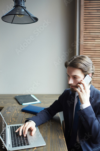 Portrait of young succesful businessman working with laptop and speaking by smartphone at wooden table