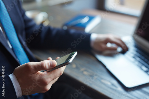 Closeup portrait of successful businessman : male hand holding smartphone, typing messages and browsing internet while working with laptop