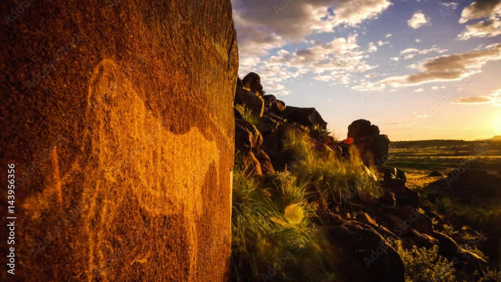 Static timelapse with a textured rock and a bushmen engraving on a hill with green grass in the foreground and the sun setting in the background with clouds moving towards the camera available on request.