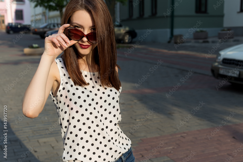 Beautiful young girl in jeans, polka-dot shirt with sunglasses walking in a sunny city Beautiful young girl in jeans, polka-dot shirt with sunglasses walking in a sunny city