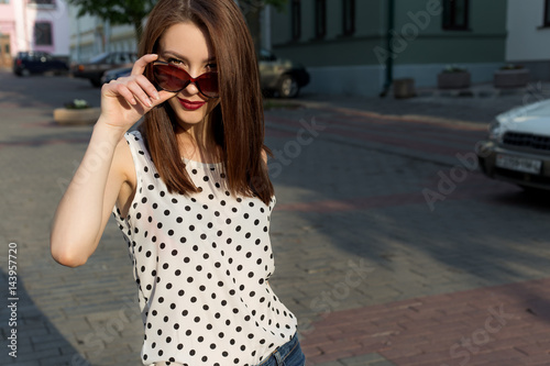 Beautiful young girl in jeans, polka-dot shirt with sunglasses walking in a sunny city