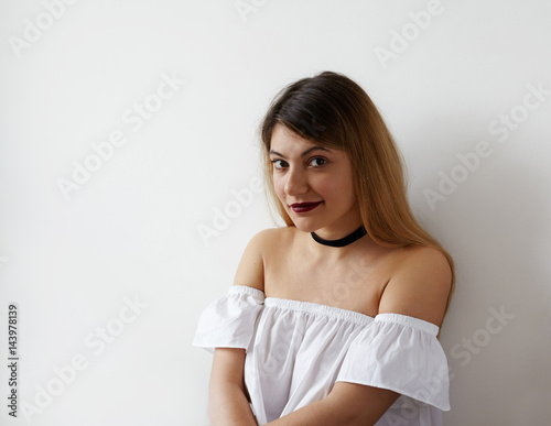 Fashionable Caucasian girl in dress with open shoulders looking at camera, posing against white wall before going out with her boyfriend, having happy and joyful look. Youth and happiness concept