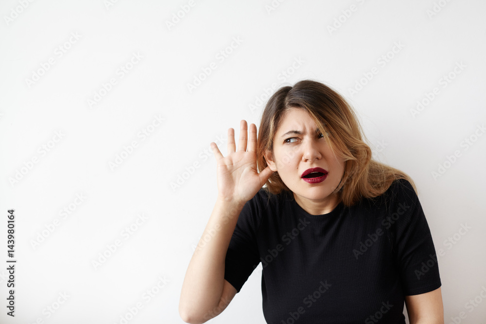 Portrait of funny plus size young woman looking aside holding hand to her ear and trying to hear something standing on white background. Cute Caucasian female with a hearing disorder or hearing loss Portrait of funny plus size young woman looking aside holding hand to her ear and trying to hear something standing on white background. Cute Caucasian female with a hearing disorder or hearing loss
