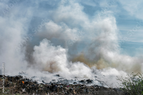 Fire at landfill with white smoke in sunny weather and clouded sky.