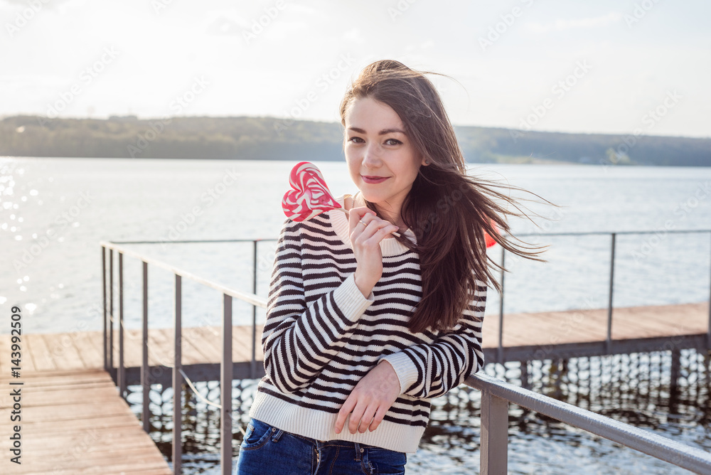 Young woman with a lollipop. Cheerful girl and a knitted hat have fun with heart shaped candy. Licking candy. Outdoors, lifestyle.