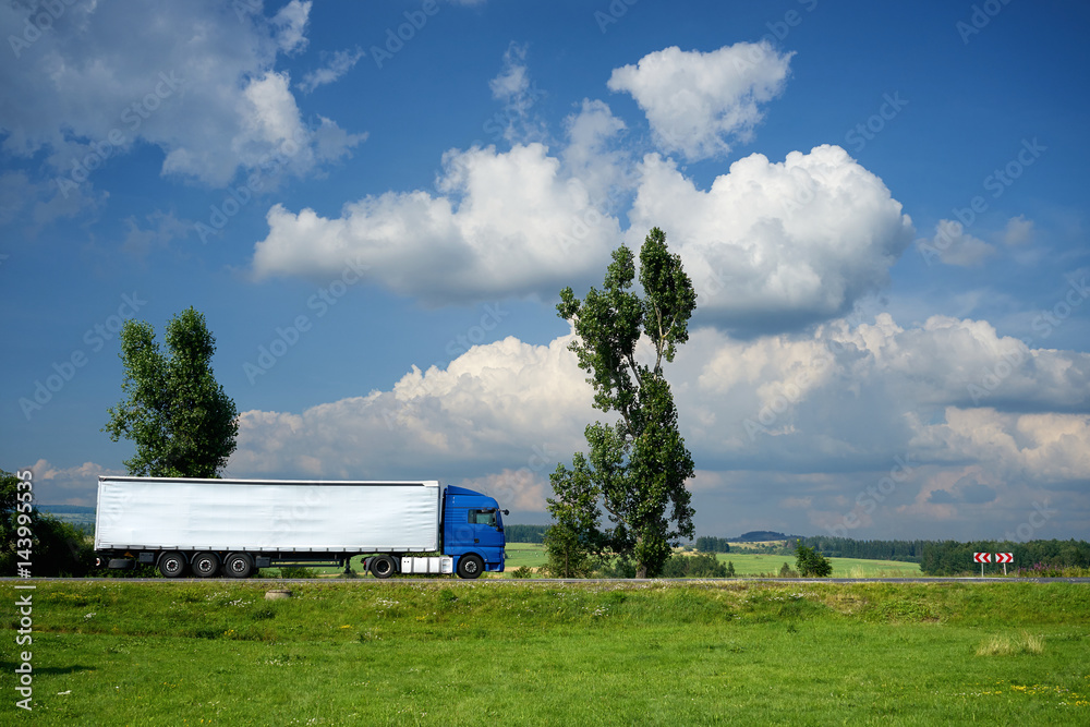 Blue truck driving on the road next to tall trees in a rural landscape under a blue sky with dramatic white clouds