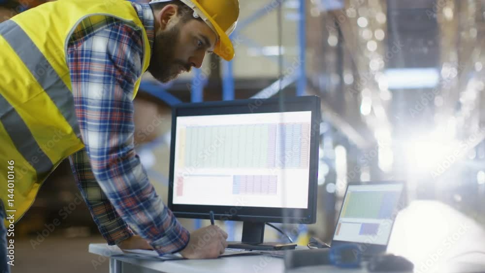 Male Inspector Fills up Spreadsheets on His Personal Computer. He's in Big Warehouse with Rows of Pallet Racks. Shot on RED EPIC-W 8K Helium Cinema Camera.