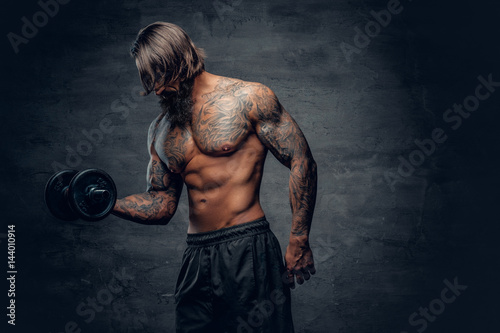 Tattooed male doing a biceps workout with dumbbell over dark grey background. © Fxquadro