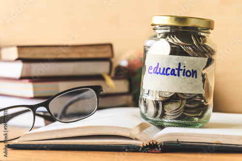 Coins in glass jar with education label, books,glasses and globe on wooden table. Financial concept.