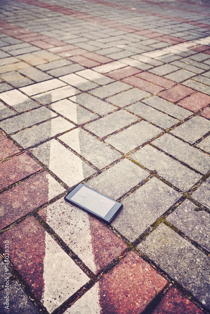 Fototapeta premium Vintage stylized picture of a mobile phone with broken screen on a parking pavement, selective focus.