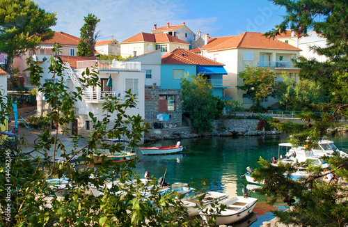 Cozy picturesque town of Bozava, Dugi otok, Croatia. Small colorful houses near emerald green water where boats and yachts are floating. Everything framed with lush green foliage.