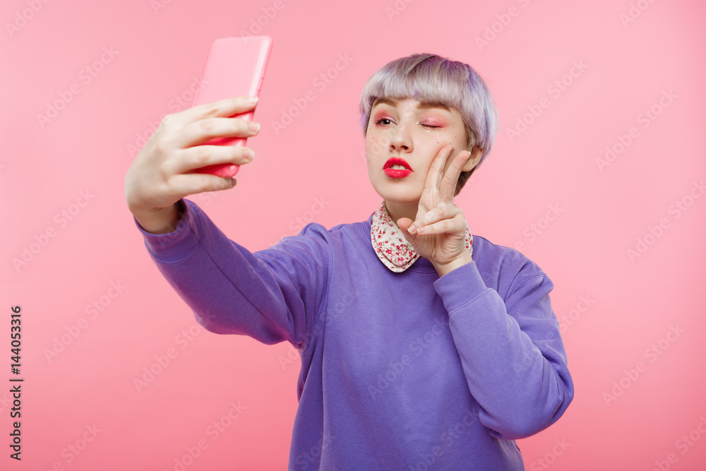 Close-up portrait of beautiful dollish girl with short light violet hair wearing lilac sweater making selfie over pink background. Close-up portrait of beautiful dollish girl with short light violet hair wearing lilac sweater making selfie over pink background.