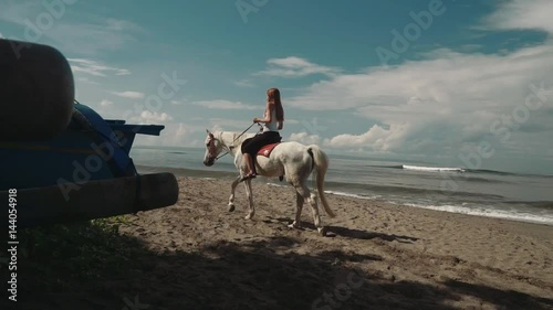 Young slim caucasian woman in white top and black leggings riding horse on beach in Bali on sunny summer day with blue sky and clouds. Moving away from camera, view from back. Shot with Sony a7s