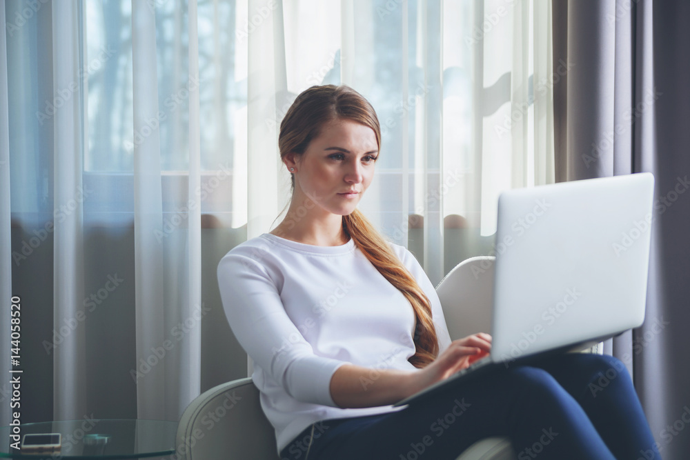 Pretty woman in modern hotel room sitting on armchair and using laptop Pretty woman in modern hotel room sitting on armchair and using laptop