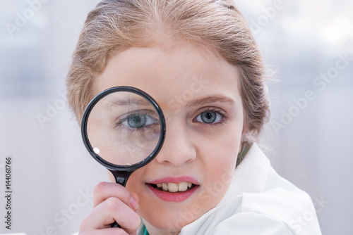 Close-up portrait of cute little girl in white coat holding magnifier looking at camera