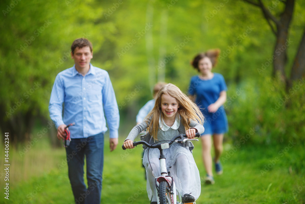 Family on a walk in the forest. A girl rushes on a bicycle on the green grass, parents try to catch up with her on foot