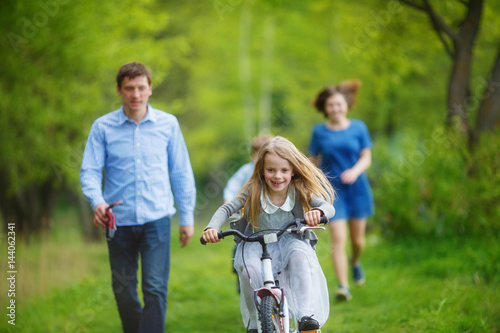 Family on a walk in the forest. A girl rushes on a bicycle on the green grass, parents try to catch up with her on foot © Daria