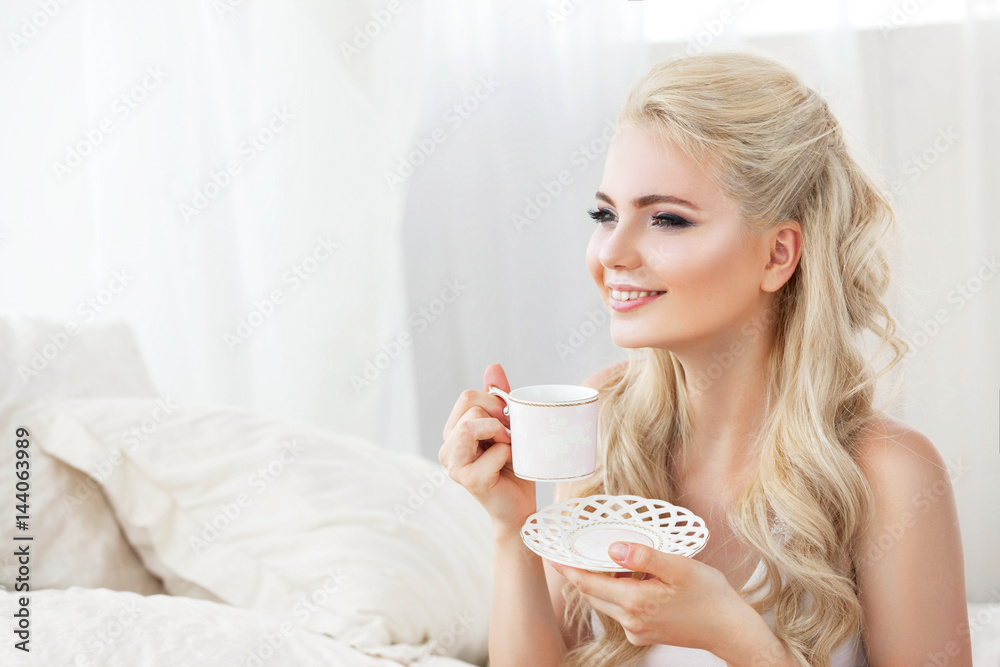 Beautiful young smiling woman sitting on white bed , holding a cup of coffee Beautiful young smiling woman sitting on white bed , holding a cup of coffee