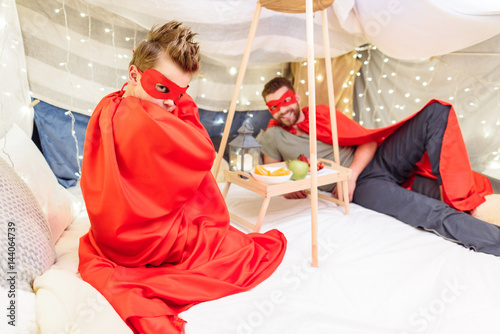 Father and son in superhero costumes playing together in blanket fort