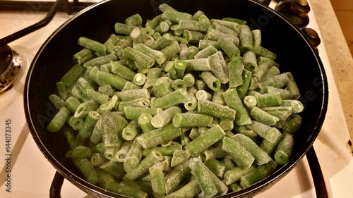 Fresh juicy green french beans are fried in a frying pan on a kitchen gas stove
