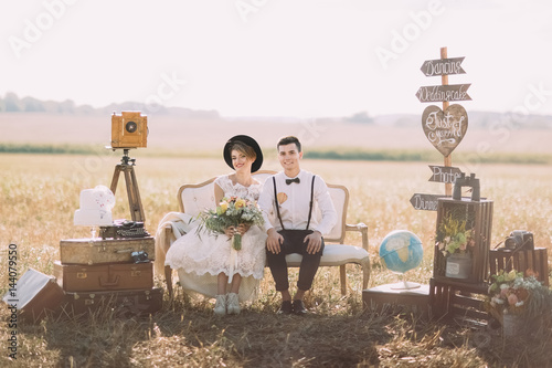 The smiling bride with bouquet and happy groom in vintage suit are sitting on the old-fashioned sofa surrounded by flowers, wooden plaques with signs, suitcases at the background of the spring field.