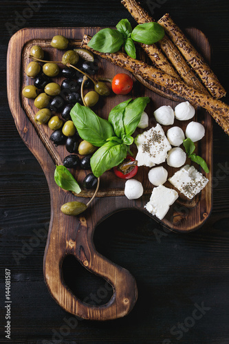 Bild auf Leinwand Mediterranean appetizer antipasti board with green black olives, feta cheese, mozzarella, capers, pepper, basil with grissini bread sticks over black wood burnt background