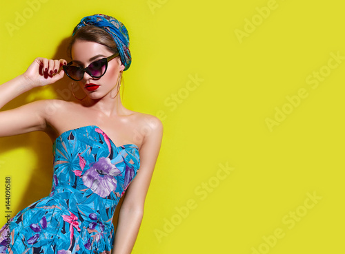 A beautiful young tanned girl with a headscarf on her head and wearing sunglasses stands near a yellow wall in the south. Fashionable blue summer dress, bright make-up, tan. photo
