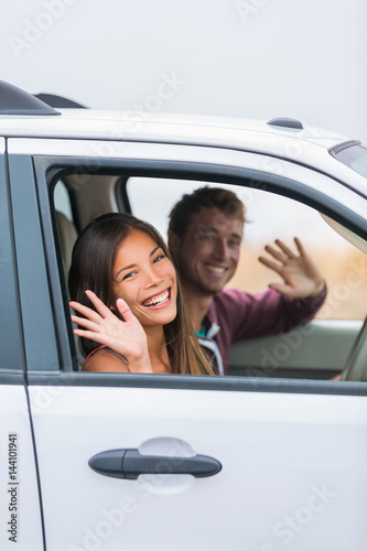 Happy multiracial couple leaving for road trip on summer vacation holiday. Young people driving car to travel destination waving goodbye. © Maridav