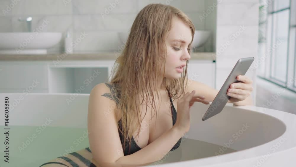 Smiling woman in black swimsuit using tablet in a bathtub Smiling woman in black swimsuit using tablet in a bathtub