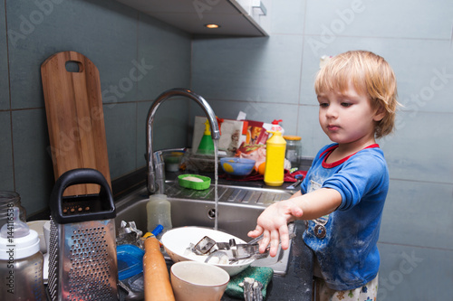 A little cute boy washing dishes in the white kitchen interior. casual lifestyle photo series in real life interior. Child with helping his parents with housework..
