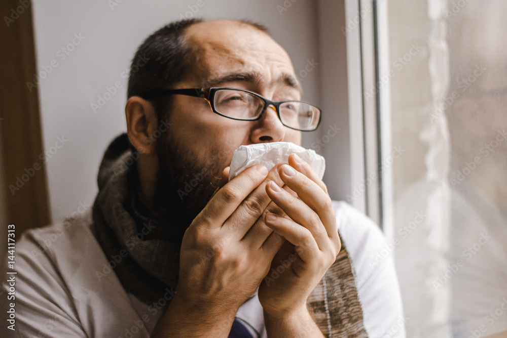 Young man blowing his nose while sitting at the window Young man blowing his nose while sitting at the window