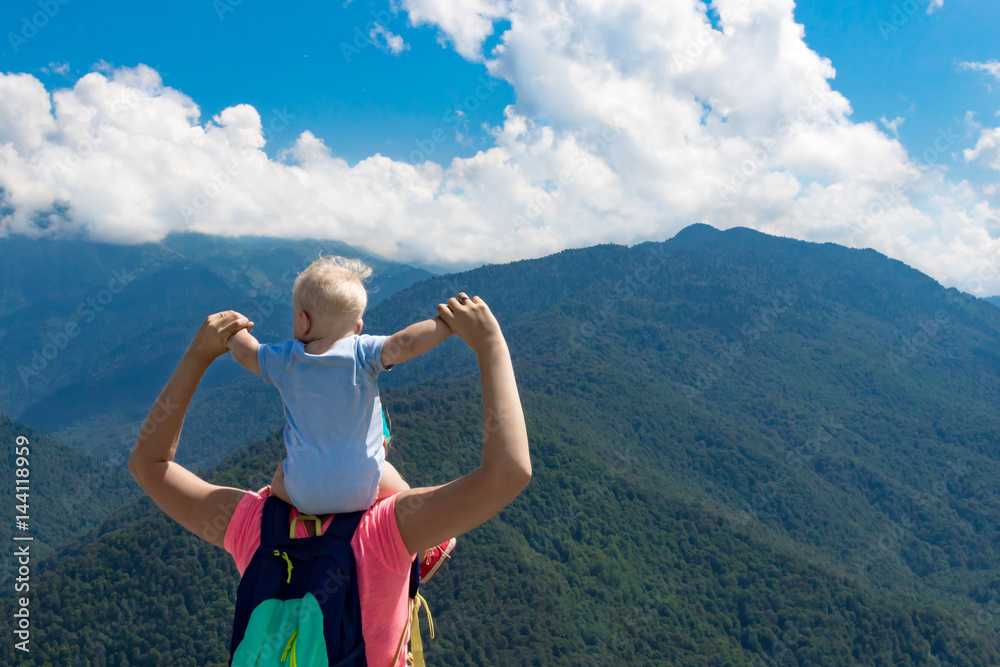 Mother or father holds his child on his shoulders near mountains and a blue sky with clouds. People look at the mountains. The parent and the child are looking at the mountains. Mother or father holds his child on his shoulders near mountains and a blue sky with clouds. People look at the mountains. The parent and the child are looking at the mountains.