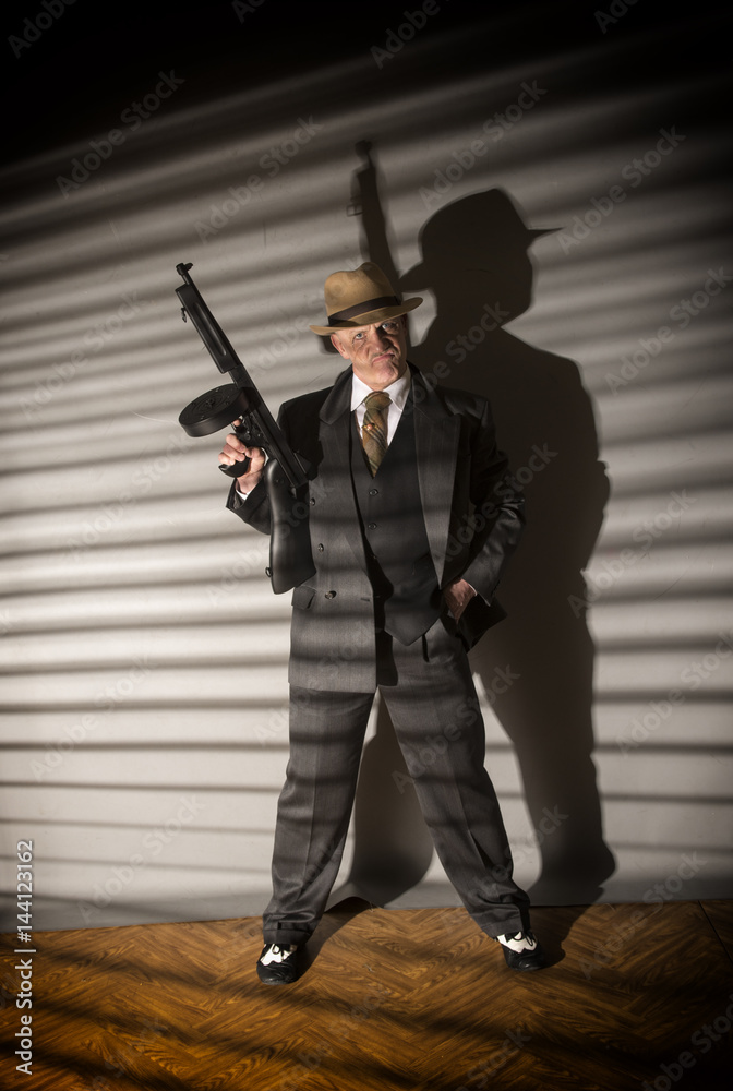Mature man dressed as a 1940s gangster holding a machine gun, on a grey background Mature man dressed as a 1940s gangster holding a machine gun, on a grey background