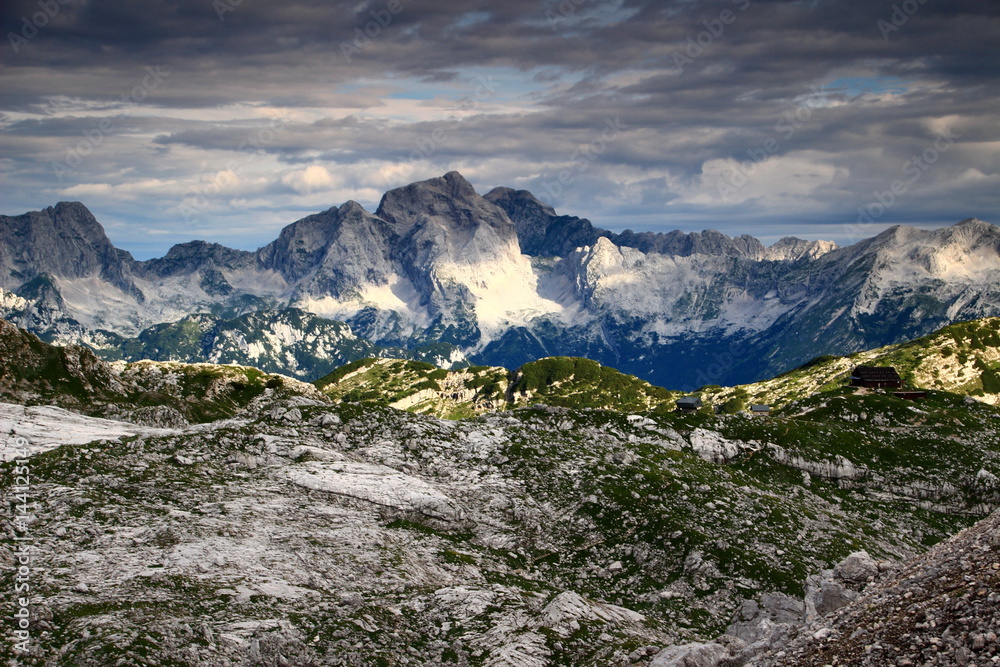 Jalovec and Mangart peaks (Julian Alps, Triglav National Park, Slovenia) in a partly sunny morning, in the foreground the Triglav Lakes Valley with grassy, rocky hills and the Prehodavci Hut.