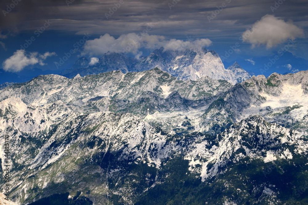 Fototapeta premium Cloud covered Jof di Montasio (Montaz) and Jof Fuart (Vis), the peaks of the Italian part of Julian Alps, rise above the Slovenian ridges in Trenta valley, seen from Kanjavec, Triglav National Park