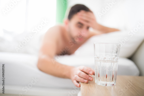 Handsome Man wake up with head pain suffers from headache and extend to get a glass of water in his bed