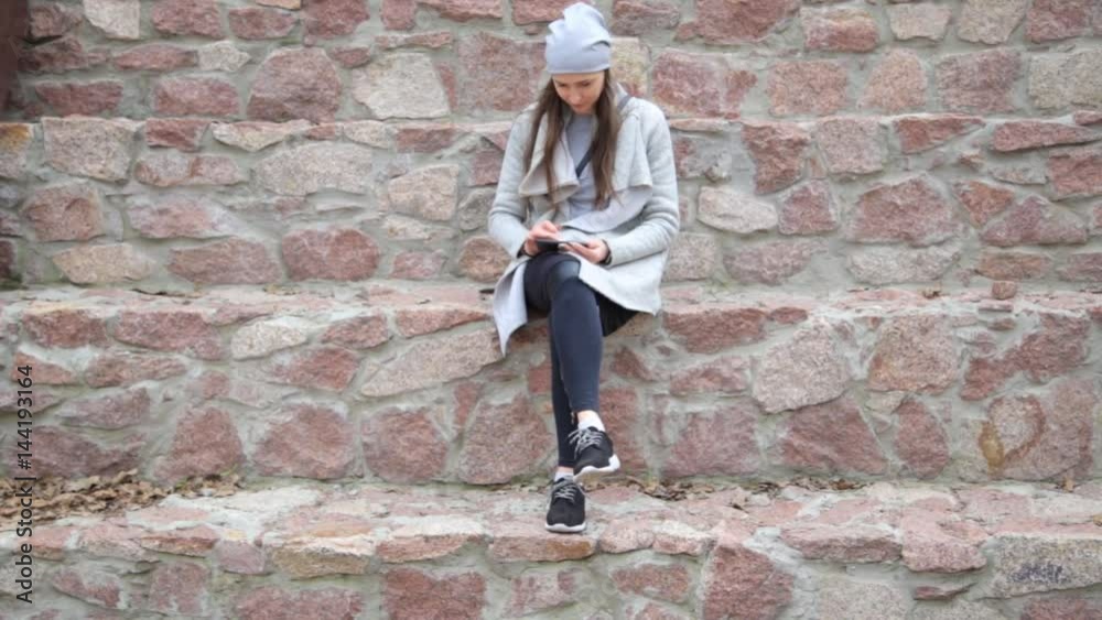 Elegant hipster woman texting on smartphone in the city. Stone wall background. Elegant hipster woman texting on smartphone in the city. Stone wall background.