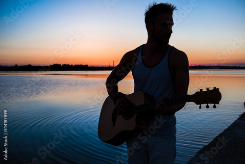 Silhouette of young handsome man playing guitar at seaside during sunrise. photo