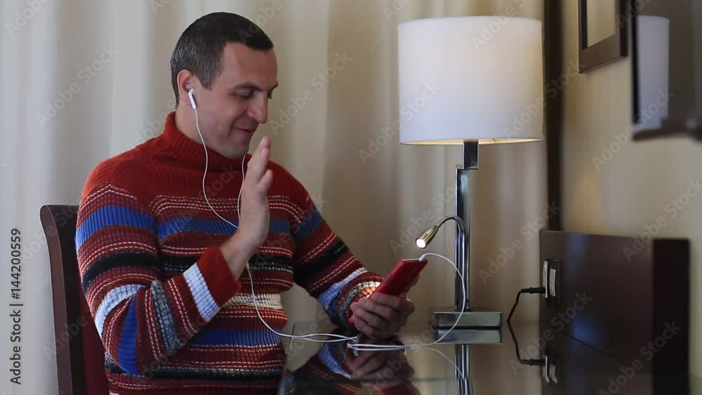 Man sits at the table in hotel room and communicates via smartphone. Cheerful man talks on cell phone. Male with smartphone communicates through skype