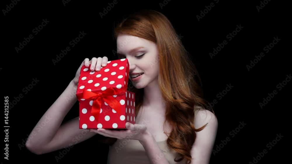 Cheerful young woman with long curly red hair looking inside present box isolated on a black background